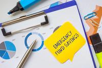The words &quot;emergency fund savings&quot; appear on a yellow sticky note that's stuck, along with a silver pen, to a set of graphs on a clipboard next to a blue highlighter.